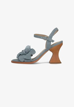 Sandalia azul de ante con correa en el tobillo, tacón de madera esculpido y flor decorativa con detalles de pétalos y pequeños abalorios en la parte frontal.