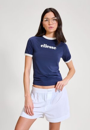 Woman with long dark hair wearing large clear glasses, navy blue ellesse t-shirt with white trim, and white elastic waist shorts.