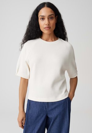 Donna con lunghi capelli ricci indossa una maglietta bianca a maniche corte e pantaloni blu, in piedi davanti a uno sfondo semplice grigio chiaro.