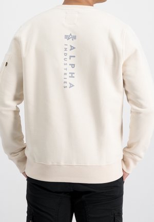 Osoba nosząca kremową bluzę z pionowym, ciemnoszarym napisem „Alpha Industries” na plecach oraz czarne spodnie bojówki, z rękami w kieszeniach.