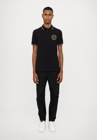 Polo negru cu accente galbene, logo pe pieptul stâng, asociat cu pantaloni negri și adidași albi. Textură netedă, ajustare clasică.
