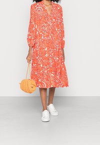 Orangenes Kleid mit floralem Muster, langen puffärmel, V-Ausschnitt und gestuften Saum. Kombiniert mit weißen Sneakers und einer runden, orangen Handtasche.