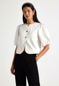 Hvid cropped bluse med korte puffærmer, der har en rund halsudskæring og tre sorte knapper ned foran. Sammen med sorte bukser.