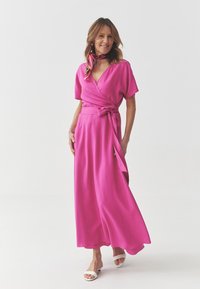 A vibrant pink wrap dress with short sleeves, a tie waist, and a flowing skirt, paired with a multi-color patterned scarf and white sandals.