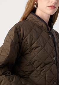 Veste brun matelassée avec un col haut, dotée de finitions noires et de boutons-pression. Le matériau a une texture lisse et légèrement réfléchissante.