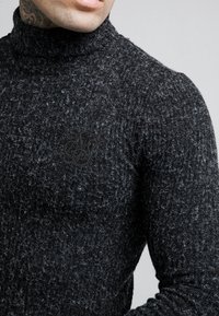 Schwarzer gerippter Rollkragenpullover mit texturiertem Finish und dezentem Logo-Detail auf der Brust. Der Stoff wirkt weich und warm.