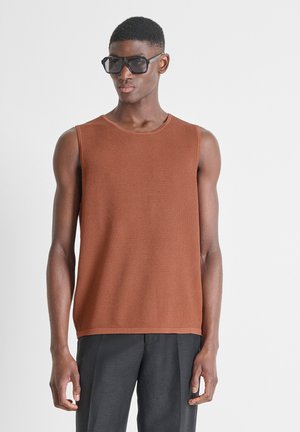 Homme portant de grandes lunettes de soleil noires, un haut marron texturé sans manches et un pantalon gris foncé, debout devant un fond blanc uni.