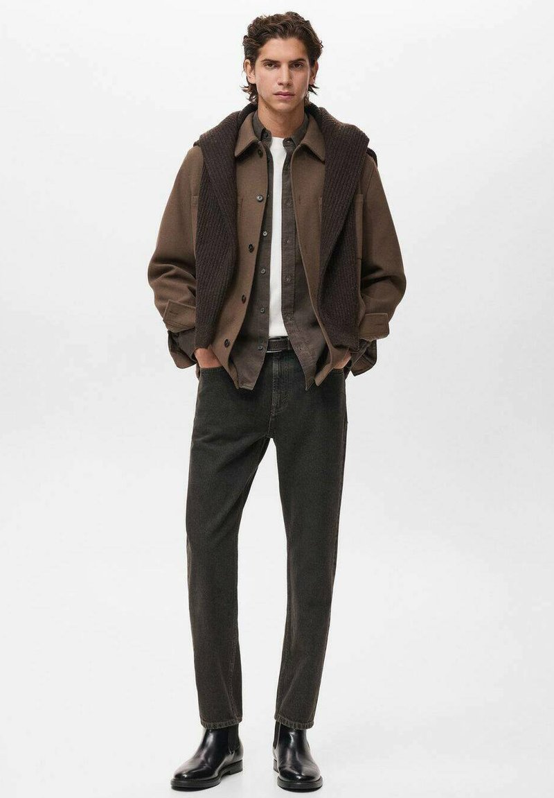 Giovane uomo che indossa una giacca marrone a strati, jeans scuri, stivali neri e un maglione appoggiato sulle spalle, in piedi con le mani nelle tasche.