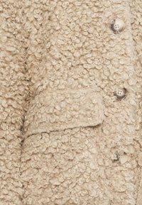 Cappotto beige dalla texture riccia con tasca applicata e bottoni chiari. Il tessuto è morbido e soffice, migliorando la sua qualità tattile.