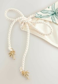 Bas de bikini couleur crème avec bordure imprimée de feuilles vertes, noué avec des cordons torsadés terminés par des breloques dorées en forme de palmier.