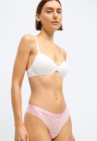 Hvit polstret bh med justerbare stropper og en rosa gjennomsiktig stringtruse med stjernemønster og logo på midjebåndet. Glatt stofftekstur.