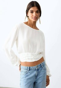 Bílý top s dlouhým rukávem a nařaseným pasem, s jemnou žebrovanou strukturou. Kombinován s světle modrými džínami.