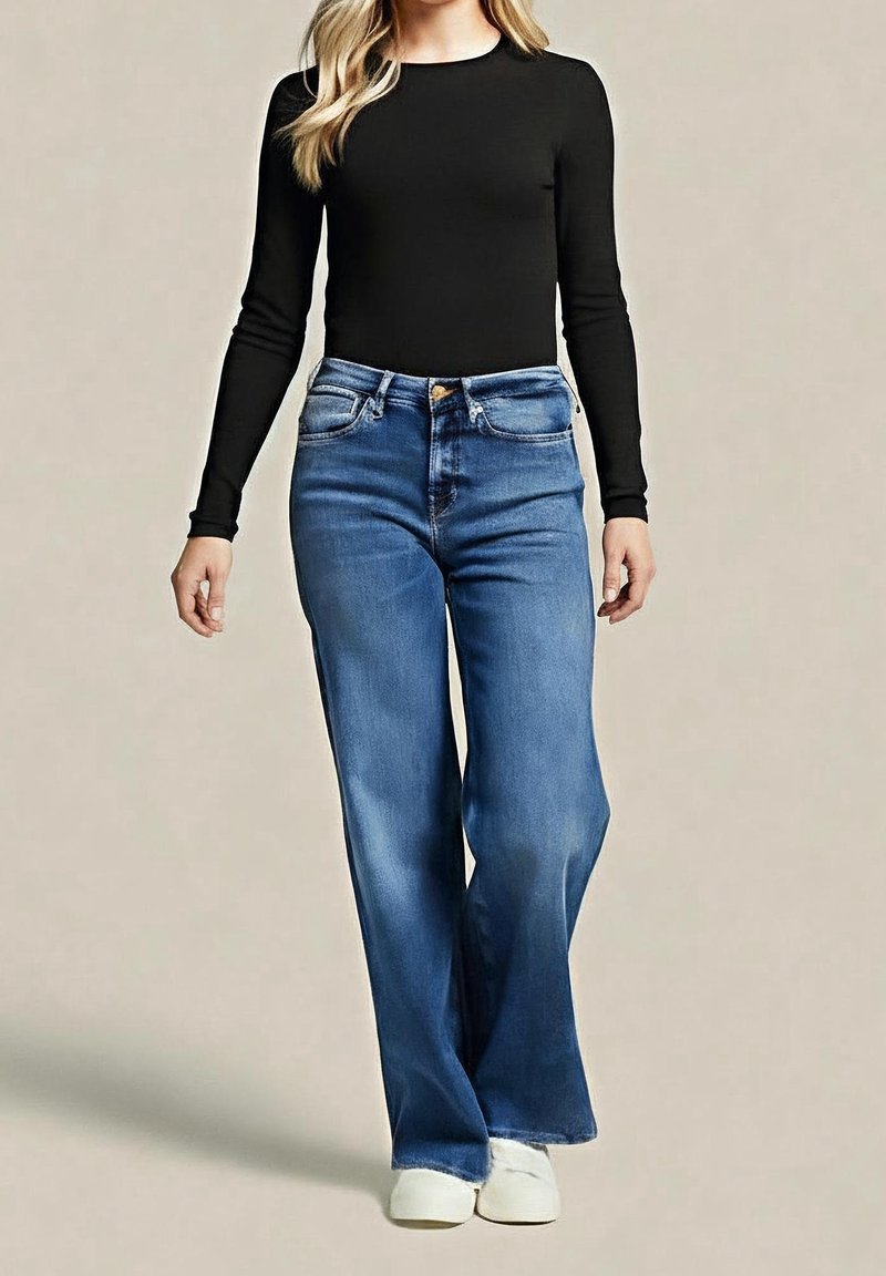 Femme portant un top noir à manches longues, un jean bleu taille haute à jambes larges et des baskets blanches, debout devant un fond uni.