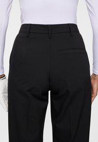 Persoon in zwarte op maat gemaakte broek en witte langetermijn top, met een witte handschoen aan de linkerhand, van achteren weergegeven.