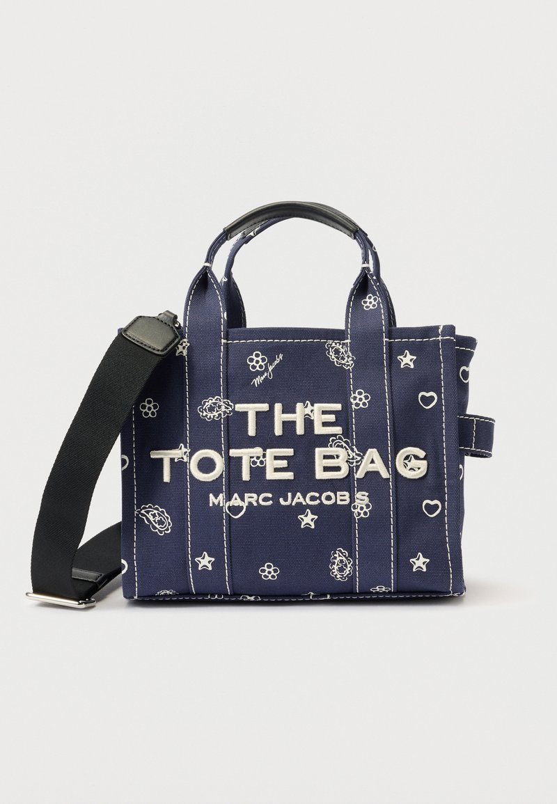 Geantă de pânză bleumarin cu imprimeuri florale și stele albe, text alb "THE TOTE BAG MARC JACOBS", curea de umăr neagră ajustabilă și mânere duble.