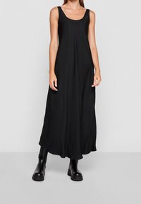 Mujer vistiendo un vestido maxi negro sin mangas combinado con botas cortas negras y gruesas, de pie contra un fondo gris claro.
