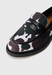 Close-up of a black and white cow print loafer with black leather strap and trim, featuring a rounded toe and low black sole.
