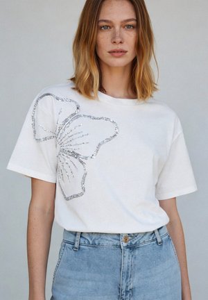 Young woman with shoulder-length hair wearing a white T-shirt with a large silver butterfly design and light blue high-waisted jeans.