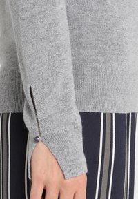 Pull en maille grise avec une finition texturée, une fente latérale et un bouton décoratif sur le poignet. Vêtement à rayures en bas partiellement visible.