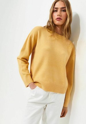 Jonge vrouw met lang blond haar, die een mosterdgele trui en witte broek draagt, staat met één hand in haar zak tegen een witte muur.
