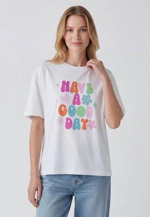 Donna con capelli biondi che indossa una t-shirt bianca con testo colorato "Have a good day" e jeans blu casual su sfondo chiaro e uniforme.