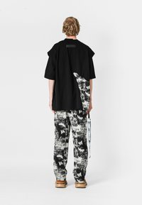 Hombre con el cabello desordenado de pie, de espaldas, vistiendo una camisa negra de gran tamaño con un estampado abstracto en blanco y pantalones a juego con un patrón en blanco y negro.