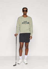 Lys grønn genser med et slottmotiv, kombinert med et svart skjørt. Modellen holder en golfkølle, iført solbriller og hvite joggesko.