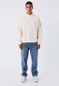 Lichtbeige polo sweatshirt met kraag, gecombineerd met blauwe denim jeans met een herhaald tekstpatroon. Draag met witte sportschoenen.