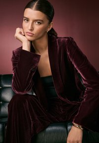 Crimson velvet suit with a tailored jacket and wide-leg pants, worn over a fitted black top, accessorized with a gold bracelet.