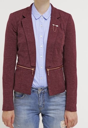 Blazer ajusté bordeaux avec poches zippées, en tissu texturé, et revers crantés, porté sur une chemise bleu clair et un jean déchiré.