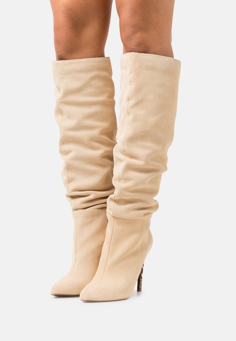 Bottes beiges jusqu'au genou en daim doux, avec un design froncé, un bout pointu et un talon aiguille haut avec un accent à motif.