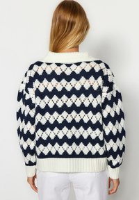 Donna che indossa un maglione lavorato a maglia a righe bianche e blu navy con colletto bianco e pantaloni bianchi, vista di spalle su uno sfondo neutro.