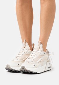 Zapatillas deportivas beige con malla texturizada y superposiciones sintéticas. Suela blanca gruesa con amortiguación de aire, logotipo negro y detalles de suela de goma.