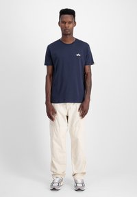 Marineblaues T-Shirt mit kurzen Ärmeln und kleinem gesticktem Logo, kombiniert mit hellen Cargo-Hosen und Turnschuhen, stehend vor einem schlichten Hintergrund.