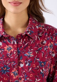 Donna che indossa una camicia a bottoni rossa con fiori blu e arancioni, visibile dalle spalle alle labbra, con capelli castani sciolti.