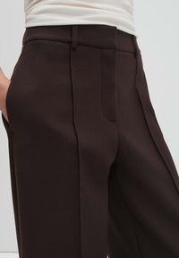 Pantaloni sartoriali marrone scuro con tasche laterali, davanti piatto e tessuto liscio e strutturato. Il design aderente valorizza la linea della vita.