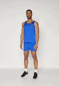 Modra športna majica brez rokavov in kratke hlače, izdelane iz lahkotnega materiala, z minimalističnim dizajnom in diskretno oznako, nošene z črnimi supergami.