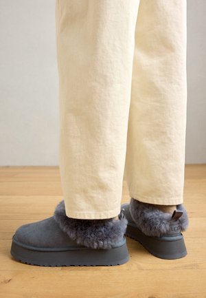 Ciabatte in suede grigio con un morbido risvolto in pelliccia grigia, suola in gomma e un piccolo dettaglio del logo sul retro. Indossate con pantaloni color crema.