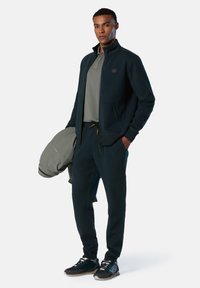 Chaqueta de cremallera azul marino oscuro con un alto cuello, combinada con una camiseta de polo gris y pantalones a juego. Las zapatillas tienen acentos en negro y azul.