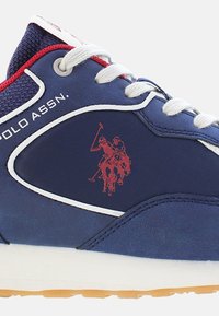 Sötétkék sneaker sima szintetikus felsőrésszel, fehér részletekkel és piros polos játékos logóval. Különböző színű fűző és textúrázott anyag a nyelvnél.