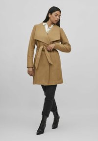 Abrigo envolvente color marrón claro con cinturón en la cintura, solapas grandes y mangas largas; combinado con jeans negros y botas de tobillo negras con punta.