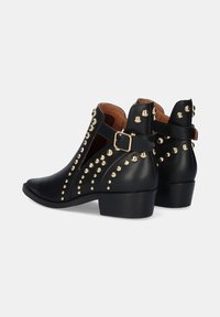 Botas de tobillo de cuero negro con tachuelas doradas, diseño de lado abierto y tacón bajo. Presenta una hebilla decorativa en el lateral. Textura suave.
