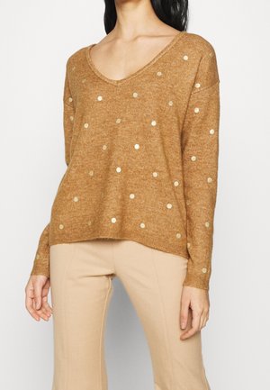 Pull en bronze avec un décolleté en V, orné de embellissements circulaires dorés ; texture douce, coupe loose, et arrière plus long. Porté avec un pantalon beige.