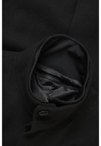 Cappotto nero con colletto alto, fodera interna liscia e dettagli con tasche a zip. Il tessuto è in lana testurizzata con accenti in bottone.