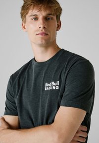 Mørkegrå kortærmet t-shirt lavet af bomuld, med et hvidt "Red Bull Racing" logo på venstre brystområde. Glat tekstur.