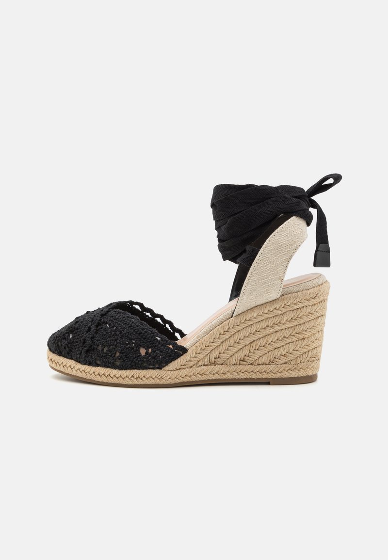 Black crocheted wedge sandal with a jute sole, featuring a beige heel counter and black fabric ankle wrap. Textured detailing throughout.