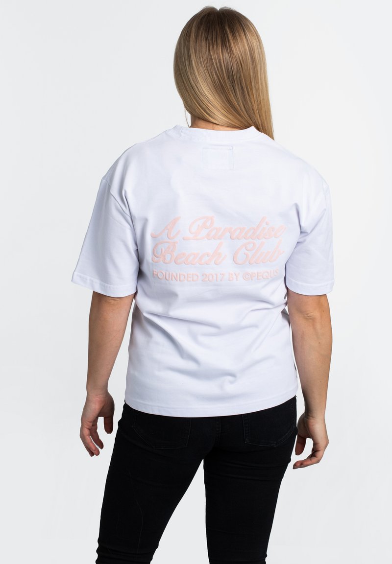 White cotton t-shirt with short sleeves, featuring pink cursive text on the back: "A Paradise Beach Club" and "Founded 2017".