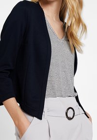 Cardigan blu navy con una finitura testurizzata, indossato sopra un top a v rigato. Pantaloni grigi chiari a vita alta con una fibbia rotonda.