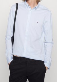 Světle modrá košile na knoflíky s límcem a dlouhými rukávy, s malým logem na hrudi, v kombinaci s černými kalhotami.