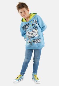 Sudadera azul claro con diseño gráfico de un oso y texto, con una capucha de un verde brillante. Combinada con jeans azules y zapatillas multicolores de caña alta.